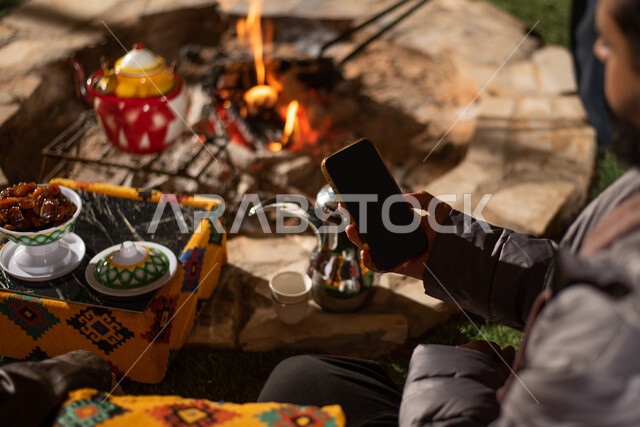 صورة مقربة لرجل خليجي عربي سعودي يجلس امام شعلة الحطب ويستخدم الهاتف المحمول، بث فيديو مباشر، استخدام وسائل التواصل الاجتماعي، مخيمات سياحية شعبية في صحاري السعودية، كشتة بر شبابية مسائية، اجواء شتوية ليلية