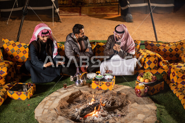 مجموعة من الاصدقاء العرب الخليجيين السعوديين يجسون اما شعلة النار بايماءات تدل على البرد الشديد، ارتداء ملابس الشتاء الدافئة، حوارات ونقاشات بين الاصدقاء، خيام شعبية في صحاري السعودية، كشتة بر شبابية مسائية، اجواء شتوية ليلية