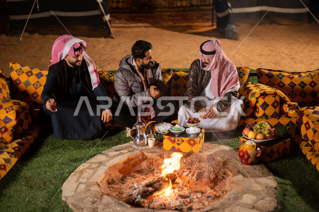 مجموعة من الاصدقاء العرب الخليجيين السعوديين يجسون اما شعلة النار بايماءات تدل على البرد الشديد، ارتداء ملابس الشتاء الدافئة، حوارات ونقاشات بين الاصدقاء، خيام شعبية في صحاري السعودية، كشتة بر شبابية مسائية، اجواء شتوية ليلية