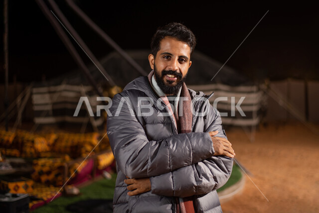 شاب خليجي عربي سعودي بأيدي متقاطعة، ارتداء ملابس الشتاء الدافئة، ايماءات وجه مبتسمة، جاكيت الشتاء، الشعور بالصقيع، مخيمات سياحية في صحاري السعودية، كشتة بر مسائية، خيام شعبية في الصحراء، اجواء شتوية ليلية