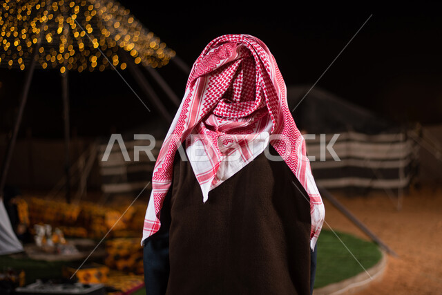 صورة من الخلف لرجل عربي خليجي سعودي يرتدي ملابس الشتاء الدافئة، جاكيت الشتاء، رحلة تخييم برية في الصحراء، مخيمات سياحية في الصحراء، خيام شعبية في صحاري السعودية، كشتة بر شبابية مسائية، اجواء شتوية ليلية