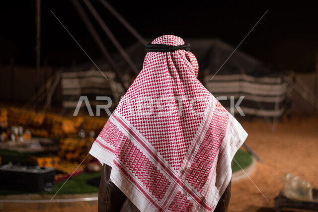 صورة من الخلف لرجل عربي خليجي سعودي يرتدي ملابس الشتاء الدافئة، جاكيت الشتاء، رحلة تخييم برية في الصحراء، مخيمات سياحية في الصحراء، خيام شعبية في صحاري السعودية، كشتة بر شبابية مسائية، اجواء شتوية ليلية