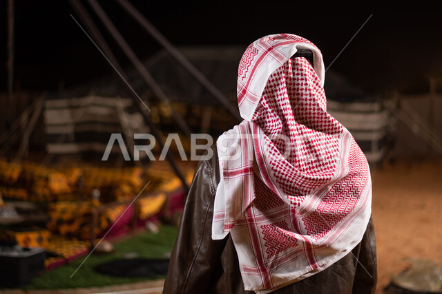 صورة من الخلف لرجل عربي خليجي سعودي يرتدي ملابس الشتاء الدافئة، جاكيت الشتاء، رحلة تخييم برية في الصحراء، مخيمات سياحية في الصحراء، خيام شعبية في صحاري السعودية، كشتة بر شبابية مسائية، اجواء شتوية ليلية
