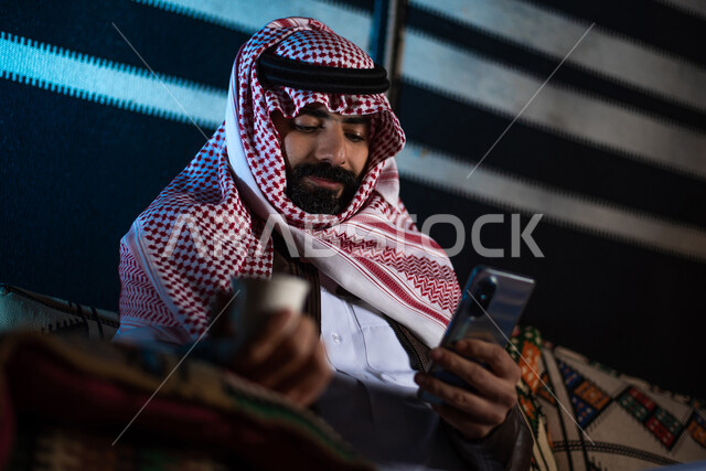 رجل عربي خليجي سعودي يرتدي ملابس الشتاء ويستخدم الهاتف المحمول، شرب القهوة الساخنة، بث فيديو مباشر، استخدام وسائل التواصل الاجتماعي، رحلة تخييم في الصحراء، جلسة بر عربية شعبية، كشتة بر شبابية مسائية، مخيمات سياحية في الصحراء، اجواء شتوية ليلية