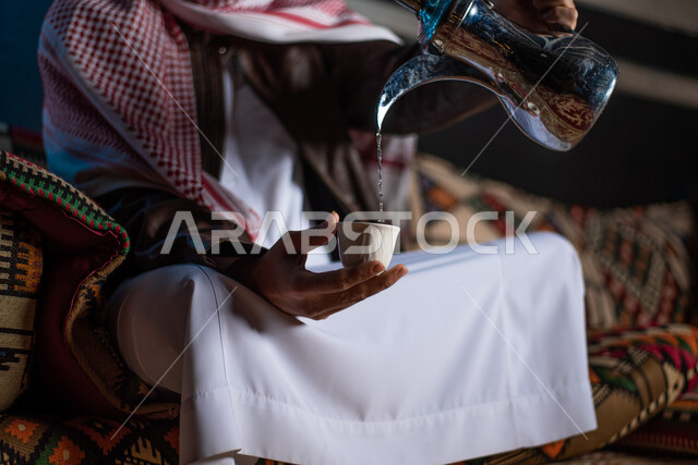 رجل خليجي سعودي عربي يرتدي ملابس الشتاء الدافئة ويقوم بصب القهوة، رحلة تخييم برية في صحراء السعودية، دلة الققهوة العربية، جلسة بر عربية شعبية، كشتة بر شبابية مسائية، مخيمات سياحية في الصحراء، اجواء شتوية ليلية