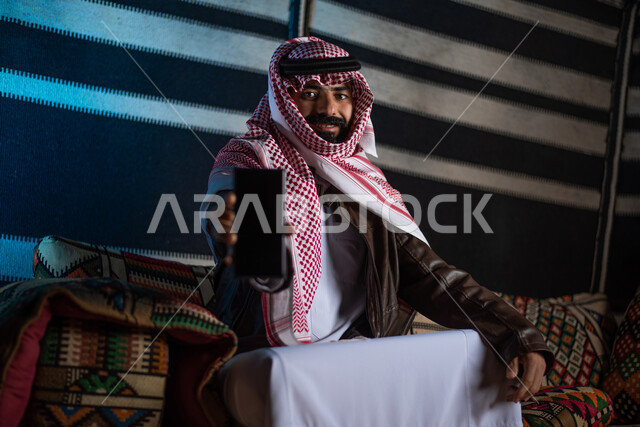 A Saudi Arab Gulf man wearing warm winter clothes, a mobile phone with a blank black screen, displaying mobile phone services and applications, electronic shopping, a popular Arab prayer session, an evening youth burial, tourist camps in the desert, a win