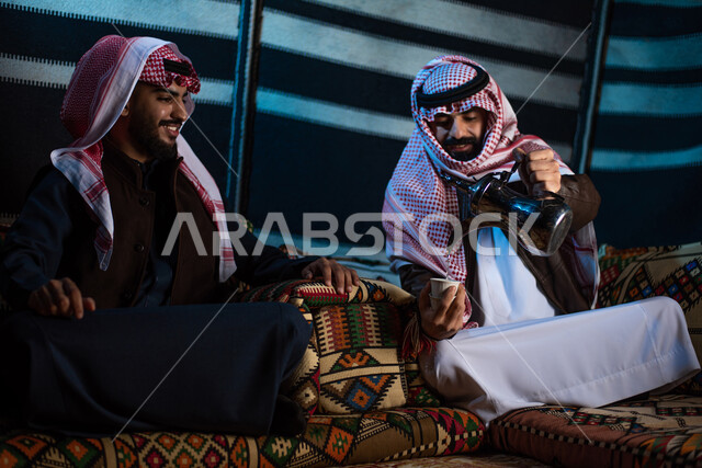 رجلان عربيان خليجيان سعوديان يستمتعان برحلة تخييم برية في صحراء السعودية، ارتداء ملابس الشتاء الدافئة، حسن الضيافة والإستقبال، تقديم القهوة الساخنة للضيوف، جلسة بر عربية شعبية، كشتة بر شبابية مسائية، مخيمات سياحية في الصحراء، اجواء شتوية ليلية