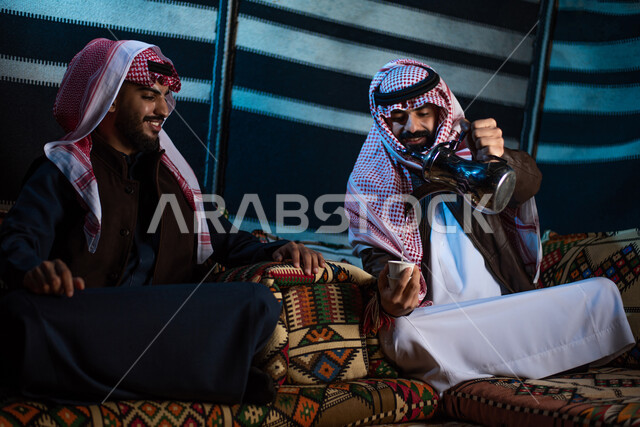 شابان عربيان خليجيان سعوديان يستمتعان برحلة تخييم برية في صحراء السعودية، ارتداء ملابس الشتاء الدافئة، حسن الضيافة والإستقبال، تقديم القهوة الساخنة للضيوف، جلسة بر عربية شعبية، كشتة بر شبابية مسائية، مخيمات سياحية في الصحراء، اجواء شتوية ليلية