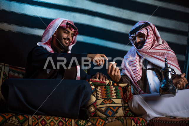 شابان عربيان خليجيان سعوديان يرتديان ملابس الشتاء الدافئة، الاستمتاع برحلة تخييم برية في صحراء السعودية، حسن الضيافة والإستقبال، تقديم القهوة الساخنة للضيوف، جلسة بر عربية شعبية، كشتة بر شبابية مسائية، مخيمات سياحية في الصحراء، اجواء شتوية ليلية