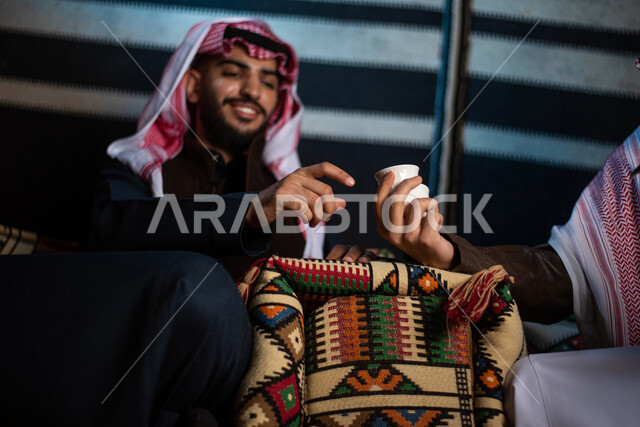 Two Saudi Arabian Gulf men enjoying a wild camping trip in the desert of Saudi Arabia, serving hot coffee to guests, enjoying drinking Arabic coffee, a popular Arabic session, an evening youth burr, tourist camps in the desert, a winter night atmosphere