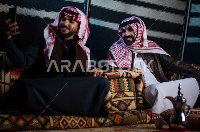 رجلان عربيان خليجيان سعوديان يستمتعان برحلة تخييم في الصحراء، ارتداء ملابس الشتاء الدافئة، التقاط صورة تذكارية من خلال الهاتف المحمول، بث فيديو مباشر، جلسة بر عربية شعبية، كشتة بر شبابية مسائية، مخيمات سياحية في الصحراء، اجواء شتوية ليلية