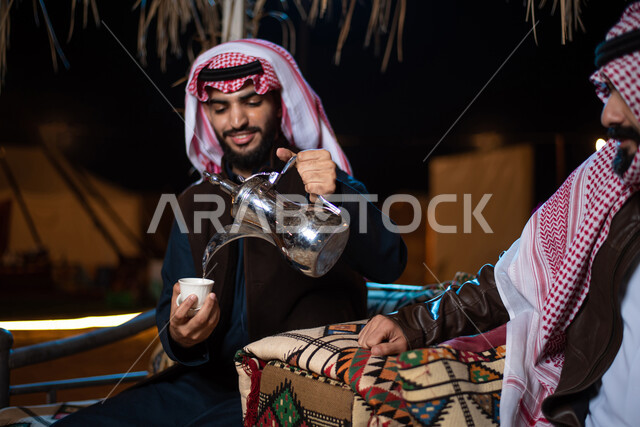 Two Saudi Arab Gulf men on a wild camping trip in the desert of Saudi Arabia, good hospitality and reception, serving hot coffee to guests, wearing warm winter clothes, a popular Arab session, an evening youth pilgrimages, tourist camps in the desert, a w