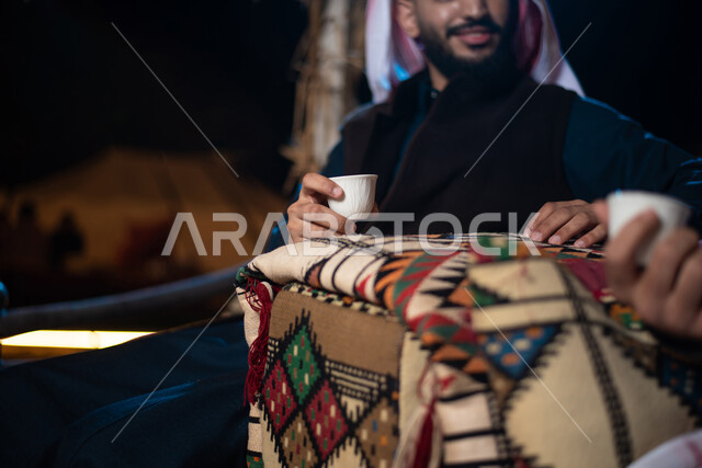 صورة مقربة لرجلان عربيان خليجيان سعوديان في رحلة تخييم برية في صحراء السعودية، تقديم القهوة الساخنة للضيوف، الاستمتاع بشرب القهوة العربية، حوارات ونقاشات بين الاصدقاء، جلسة بر عربية شعبية، كشتة بر شبابية مسائية، مخيمات سياحية في الصحراء، اجواء شتوية ليلية