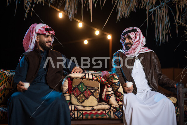 شابان عربيان خليجيان سعوديان يستمتعان بشرب القهوة العربية، رحلة تخييم برية  في صحراء السعودية، تقديم القهوة الساخنة للضيوف، حوارات ونقاشات بين الاصدقاء، جلسة بر عربية شعبية، كشتة بر شبابية مسائية، مخيمات سياحية في الصحراء، اجواء شتوية ليلية