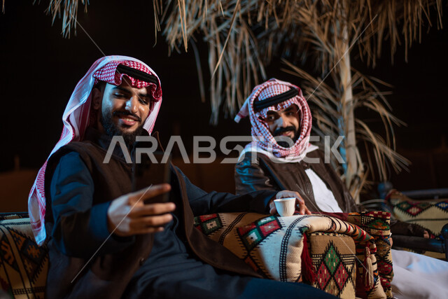 رجلان عربيان خليجيان سعوديان يرتديان ملابس الشتاء الدافئة، التقاط صورة تذكارية من خلال الهاتف المحمول، بث فيديو مباشر، رحلة تخييم في الصحراء، جلسة بر عربية شعبية، كشتة بر شبابية مسائية، مخيمات سياحية في الصحراء، اجواء شتوية ليلية