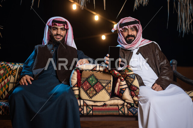 شابان عربيان خليجيان سعوديان يرتديان ملابس الشتاء الدافئة، هاتف محمول بشاشة سوداء فارغة، عرض خدمات وتطبيقات الهاتف المحمول، التسوق الإلكتروني، جلسة بر عربية شعبية، كشتة بر شبابية مسائية، مخيمات سياحية في الصحراء، اجواء شتوية ليلية
