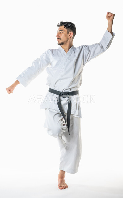 التدرب على حركات قتالية خاصة, بورتريه  لرجل عربي خليجي سعودي يمارس رياضة الكاراتيه اليابانية، تطبيق حركات الكاتا, زي الكاراتيه الأبيض مع الحزام الأسود، فنون الدفاع عن النفس ، فنون قتالية يابانية، صورة الجسم بالطول الكامل, خلفية بيضاء
