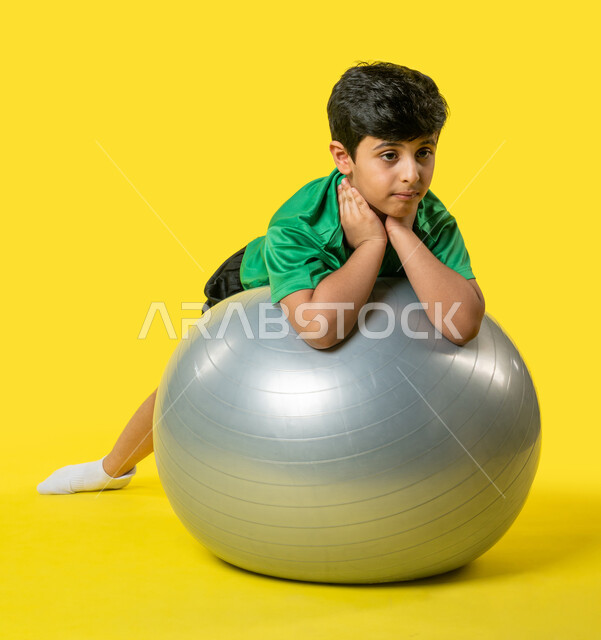 Portrait of a Saudi Arabian Gulf boy wearing sports clothes, exercising, using a Pilates ball, fitness and balance ball, children's sports activities, health and fitness, yellow background