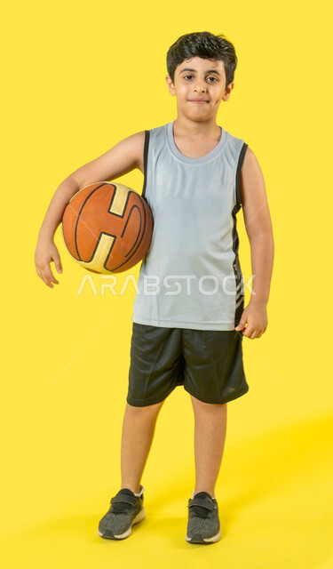 ايماءات تدل على السعادة, بورتريه لولد عربي خليجي سعودي يرتدي الملابس الرياضية يحمل تحت ذراعه كرة السلة, علامة الجودة,  هواية اللعب بكرة السلة, انشطة رياضية ترفيهية للاطفال, صورة الجسم بالطول الكامل, خلفية صفراء