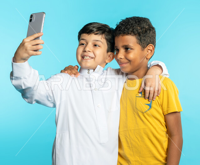 Portrait of two Saudi Arabian Gulf children using a mobile phone, two friends taking a selfie, a group photo, taking souvenir photos, documenting happy moments, making a video call, live broadcast, face and hand gestures indicating happiness, blue backgro