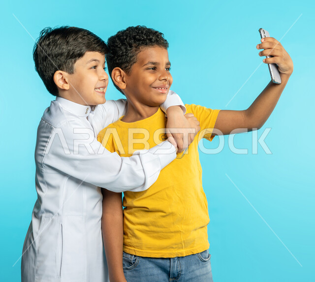 Portrait of two Saudi Arabian Gulf children using a mobile phone, two friends taking souvenir photos, taking a selfie, group photo, documenting happy moments, making a video call, live broadcast, face and hand gestures indicating happiness, blue backgroun