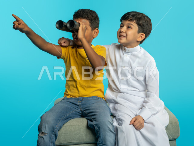 بورتريه لطفلان عربيان خليجيان سعوديان ينظران من خلال المنظار ثنائي العينين (التلسكوب)، الاشارة نحو شيء ما، الاستكشاف والمغامرة، فريق الكشافة، رؤية مستقبلية، التخطيط المستقبلي، رؤية الاشياء البعيدة، خلفية زرقاء