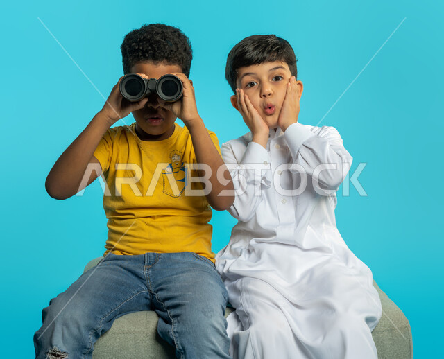بورتريه لطفلان عربيان خليجيان سعوديان ينظران من خلال المنظار ثنائي العينين (التلسكوب)، الاستكشاف والمغامرة، فريق الكشافة، رؤية مستقبلية، التخطيط المستقبلي، رؤية الاشياء البعيدة، ايماءات وجه ويدين تدل على الدهشة والصدمة، خلفية زرقاء
