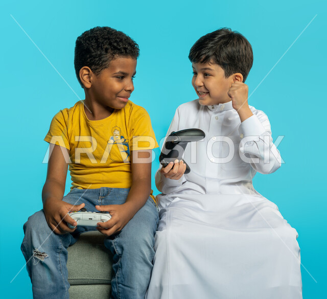 بورتريه لطفلان عربيان خليجيان سعوديان يحملان عصا التحكم، صديقان يستمتعان بالعاب الفيديو، اللعب والترفيه، التحدي والمغامرة، مفهوم الفوز والخسارة، تقنيات وألعاب، ايماءات وجه ويدين تدل على الفوز والانتصار، خلفية زرقاء