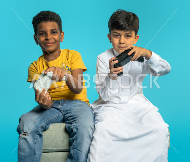 بورتريه لطفلان عربيان خليجيان سعوديان يحملان عصا التحكم، صديقان يلعبان العاب الفيديو، اللعب والترفيه، التحدي والمغامرة، مفهوم الفوز والخسارة، تقنيات وألعاب، خلفية زرقاء
