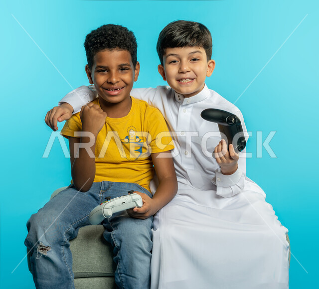 بورتريه لطفلان عربيان خليجيان سعوديان يحملان عصا التحكم، صديقان يستمتعان بلعب العاب الفيديو، اللعب والترفيه، التحدي والمغامرة، مفهوم الفوز والخسارة، تقنيات وألعاب، ايماءات وجه ويدين تدل على الفوز والانتصار، خلفية زرقاء
