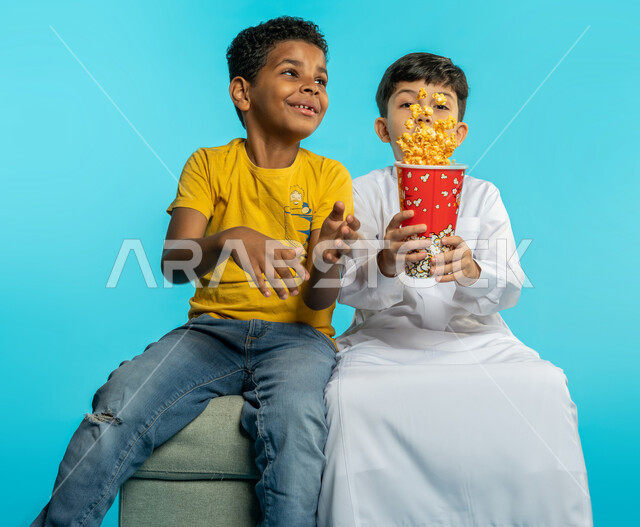 Portrait of two Saudi Arabian Gulf children with different face and hand gestures, flying popcorn grains, eating popcorn, enjoying watching movies, watching a movie, entertainment and leisure, spending enjoyable times, astonishment and amazement, blue bac