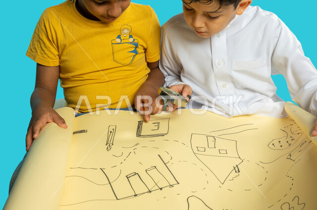 صورة مقربة لطفلان عربيان خليجيان سعوديان يستخدمان العدسة المكبرة في فحص خريطة ورقية، مفهوم الفحص والإستكشاف، الرؤية عن قرب، التحقق والتدقيق، تكبير الاشياء، أدوات بصرية، خلفية زرقاء