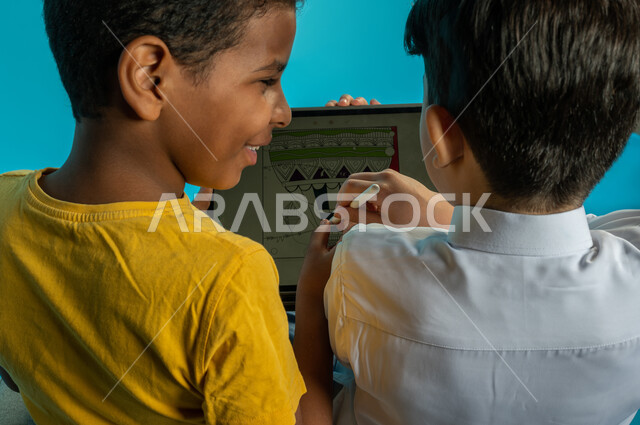 صورة مقربة من الخلف لطفلان عربيان خليجيان سعوديان يستخدمان التابليت المحمول، جهاز لوحي، استخدام قلم التابليت، الاستمتاع بمشاهدة البرامج الترفيهية من خلال التابليت، صداقة اطفال، خلفية زرقاء