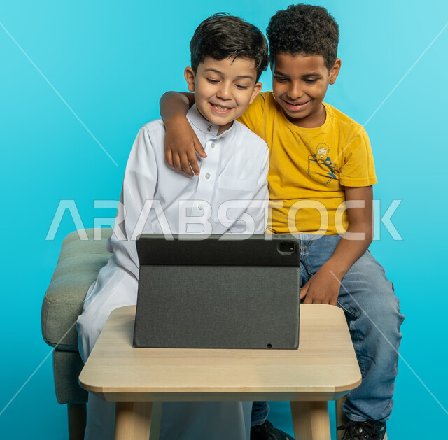 Portrait of two Saudi Arabian Gulf children, using a tablet, enjoying watching entertainment programs through a portable tablet, children's friendship, happy facial gestures, blue background