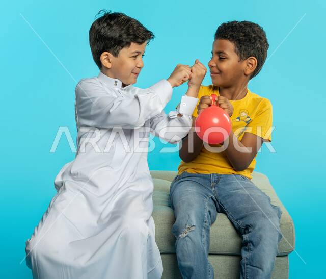 بورتريه لطفلان عربيان خليجيان سعوديان، نفخ البلالين، اللعب والمرح، بالونات ملونة، صداقة اطفال، انشطة ترفيهية، ايماءات وجه تدل على السعادة، خلفية زرقاء