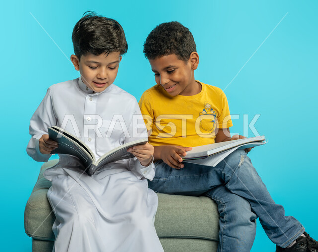 بورتريه لطفلان عربيان خليجيان سعوديان يستمتعان بالقراءة، قراءة الكتب، كتاب مدرسي، قصص وروايات، اكتساب المعرفة، تعليم وثقافة، خلفية زرقاء