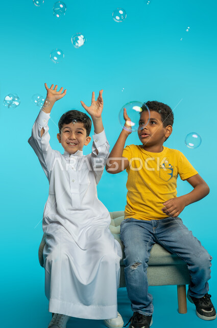 Portrait of two Saudi Arabian Gulf children catching water bubbles, playing with water bubbles, entertainment activities, children's friendship, face and hand gestures indicating happiness, blue background