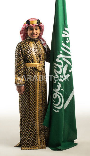 Portrait of a Saudi Arabian Gulf boy, standing next to the Saudi flag, wearing the traditional Saudi folk dress, national celebrations in the central region, the identity of the Saudi National Day 92, the Saudi founding day, a Saudi traditional costume fo