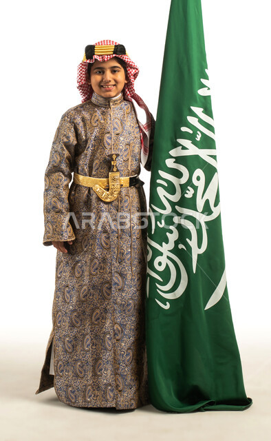 بورتريه لولد عربي خليجي سعودي، يقف بجانب علم السعودية، إرتداء الزي السعودي الشعبي التقليدي، الإحتفالات الوطنية في المنطقة الوسطى، هوية اليوم الوطني السعودي 92، يوم التأسيس السعودي، زي تراثي سعودي للمناسبات الوطنية، العيد الوطني، صورة بالطول الكامل