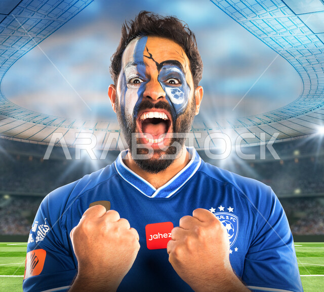 شاب عربي خليجي سعودي يرتدي تيشرت نادي الهلال السعودي، تشجيع فريق الهلال السعودي لكرة قدم، متابعة مباراة كرة قدم، شاب يرسم على وجهه شعار نادي الهلال السعودي، إيماءات وجه ويدين تدل على السعادة والحماس والشغف بالفوز