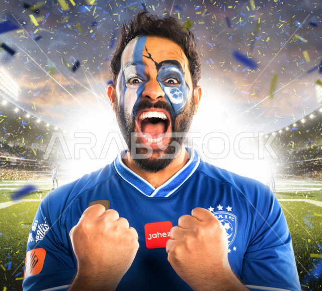 شاب عربي خليجي سعودي يرتدي تيشرت نادي الهلال السعودي، تشجيع فريق الهلال السعودي لكرة قدم، متابعة مباراة كرة قدم، شاب يرسم على وجهه شعار نادي الهلال السعودي، إيماءات وجه ويدين تدل على السعادة والحماس والشغف بالفوز