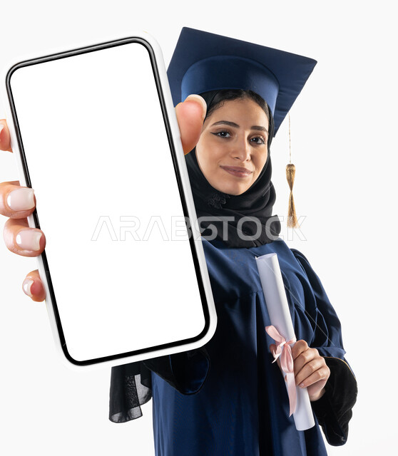 بورتريه لطالبة عربية خليجية سعودية ترتدي ملابس التخرج وتمسك بيدها شهادة النجاح والتفوق، ايماءات وجه تدل على السعادة، هاتف محمول كبير الحجم، شاشة بيضاء فارغة، خريجة جامعية، حفل تخرج، تلميذة متفوقة، مدارس وجامعات، التعليم في السعودية، خلفية بيضاء