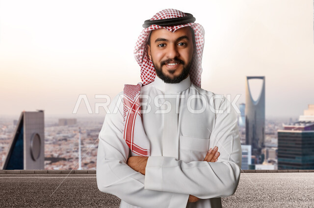صورة مقربة لرجل عربي خليجي سعودي بايدي متقاطعة، الابراج وناطحات السحاب، ايماءات وجه مبتسمة، الوقوف بثقة، خلفية ابراج الرياض