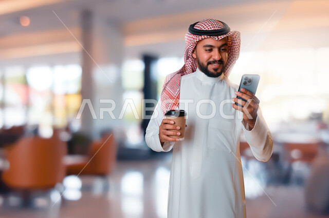 شاب عربي خليجي سعودي يستخدم الهاتف المحمول وبيده كوب، الاستمتاع بالمشروبات الساخنة، التسوق الالكتروني، طلبات اونلاين، التسوق الذكي، خدمات التوصيل السريع للمشروبات الساخنة، تصفح المواقع الالكترونية