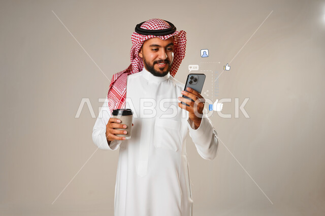 بورتريه لشاب عربي خليجي سعودي يستخدم الهاتف المحمول وبيده كوب، الاستمتاع بالمشروبات الساخنة، تصفح الفيس بوك، ايقونات ورموز الفيسبوك، المشروبات الساخنة، تصفح المواقع الالكترونية، خلفية رمادية