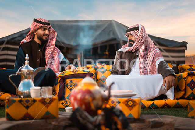 رجلان عربيان خليجيان سعوديان يرتديان ملابس الشتاء الدافئة، دلة قهوة عربية، حسن الضيافه والإستقبال، حوارات ونقاشات، صداقات وتعارف، اماكن تخييم، رحلة تخييم برية، كشتة بر، اجواء شتوية، مخيمات سياحية في الصحراء، خيام شعبية في صحاري السعودية