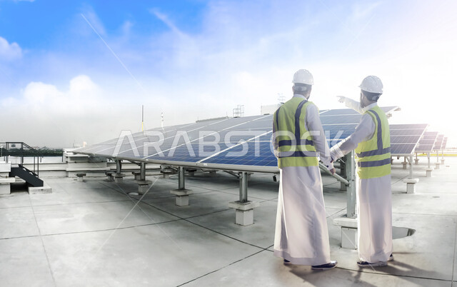 مجموعة من ألواح وأنظمة  الطاقة الشمسية ، مهندسان عربيان خليجيان سعوديان يشرفان على محطات الطاقة الشمسية ، إرتداء الزي السعودي التقليدي و سترة الحماية والخوذة  ، الإشراف على العمل، محطات توليد الطاقة المتجددة , خلفية السماء  .