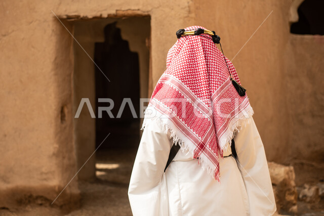 تصوير من الخلف لرجل عربي سعودي خليجي يرتدي الزي السعودي الشعبي التقليدي, الزي التراثي للمناسبات الوطنيه, التجول في إحدى المباني الطينيه الشعبيه, بيوت تراثيه قديمه, شعبيات, أحياء أثريه قديمه, جوله بين الأحياء الشعبيه