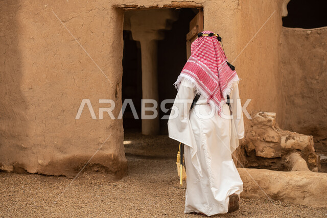 Photography from the back of a Saudi Arabian Gulf man wearing the traditional Saudi folk costume, the traditional costume for national occasions, touring in one of the popular mud buildings, old heritage houses, folklore, ancient ancient neighborhoods, a 
