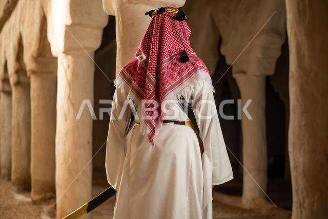 تصوير من الخلف لرجل عربي سعودي خليجي يرتدي الزي السعودي الشعبي التقليدي, الزي التراثي للمناسبات الوطنيه, التجول في إحدى المباني الطينيه الشعبيه, بيوت تراثيه قديمه, شعبيات, أحياء أثريه قديمه, جوله بين الأحياء الشعبيه